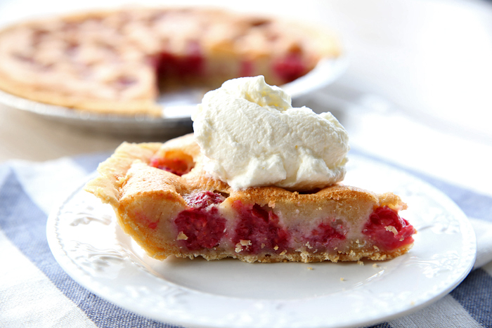 A slice of brown butter raspberry pie with a dollop of whipped cream sitting on a small plate with a striped placemat underneath. Faded in the background is a Brown Butter Raspberry Pie with a slice cut out of it.