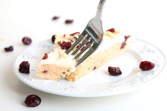 A piece of cranberry bliss bars on a white plate with a fork about to take a bite. Extra craisins on and around the plate for decoration. 