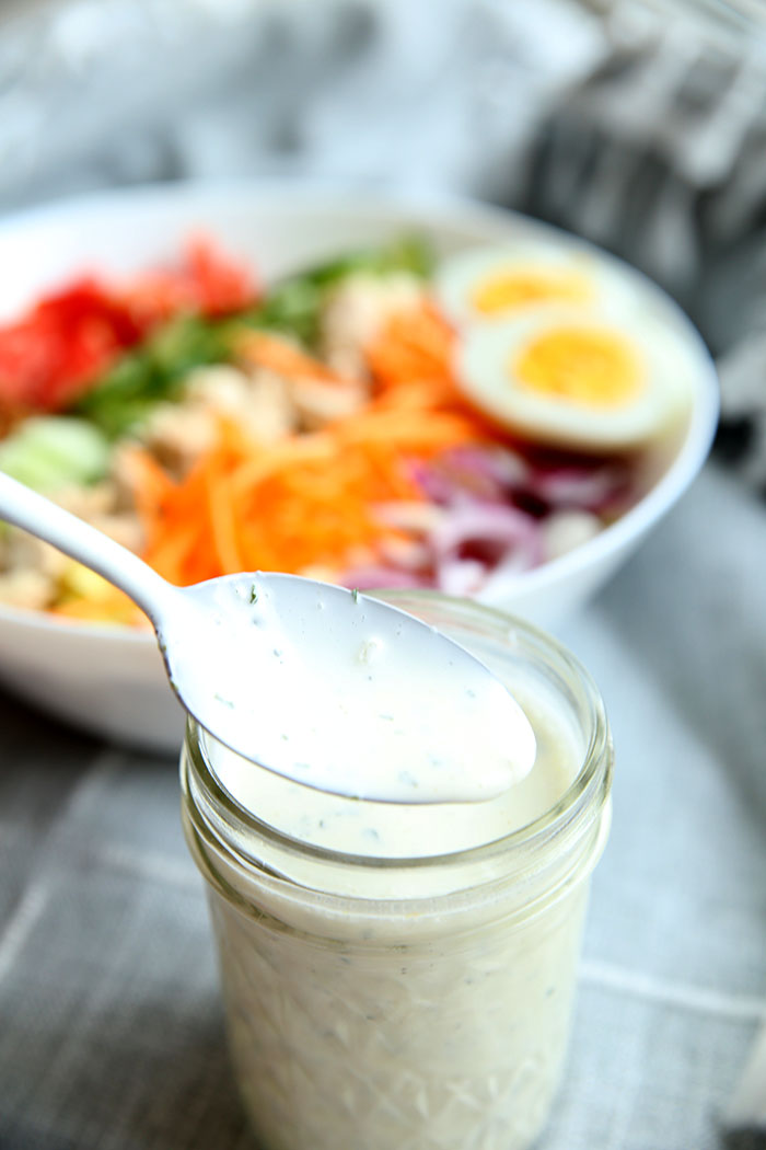 Glass jar full of homemade ranch dressing with a spoon taking some out. There is a large bowl of salad in the background