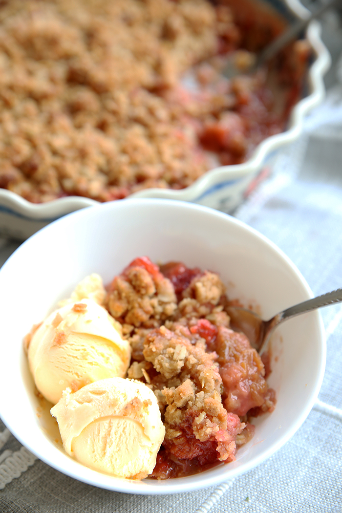 White bowl with strawberry rhubarb crisp in it and two scoops of vanilla icecream, in the background you can see a polish pottery pie dish with the rest of the crisp in it waiting to be served