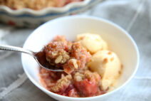 strawberry rhubarb crisp in a white bowl with a spoon taking a bite out of it. there is also vanilla icecream in the bowl, you can see a small piece of the polish pottery pie dish the rest of the crisp is in, in the background.