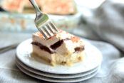 a piece of a four layer chocolate lasagna with a shortbread crust sitting on a stack of three small white plates. a fork is taking a bite off of the square of dessert. You can see the rest of the pan of dessert in the background.