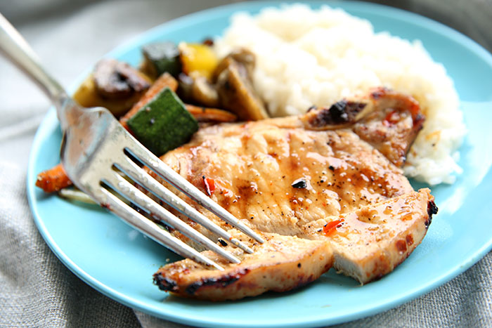 pork chop on a blue plate with coconut rice and grilled veggies in the back ground, a fork is about to take a piece that has been cut for a bite