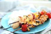three grilled teriyaki chicken kabobs on a blue palte with a side of coconut rice. Sitting on a grey and white table cloth