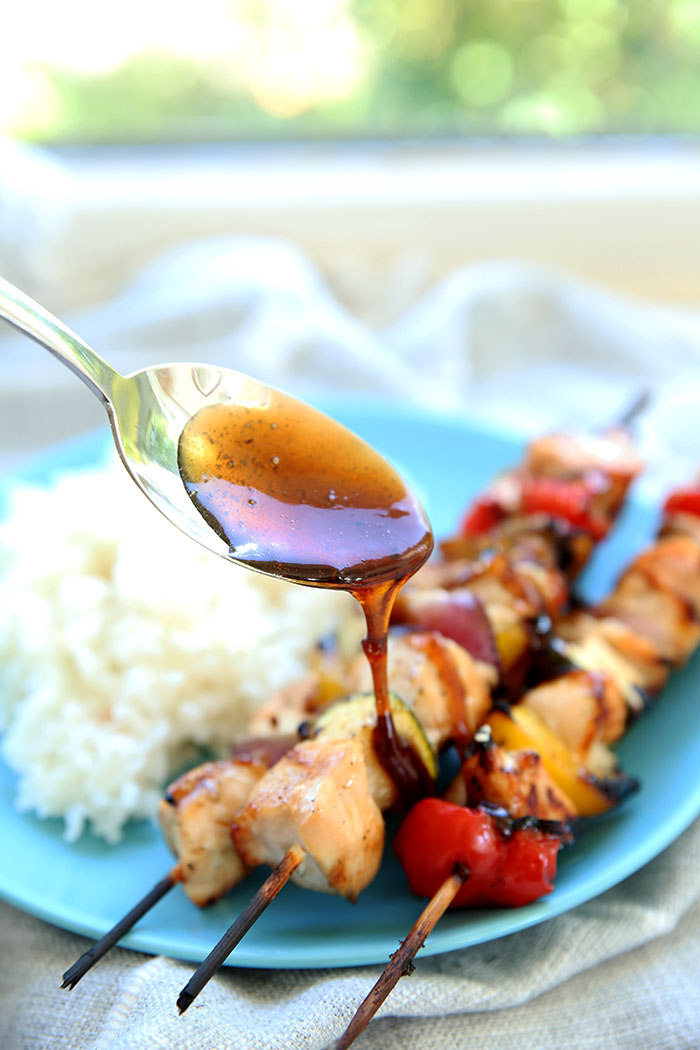 Three grilled chicken kabobs being drizzled with teriyaki sauce sitting on a blue plate next to some coconut rice that has been drizzled with teriyaki sauce