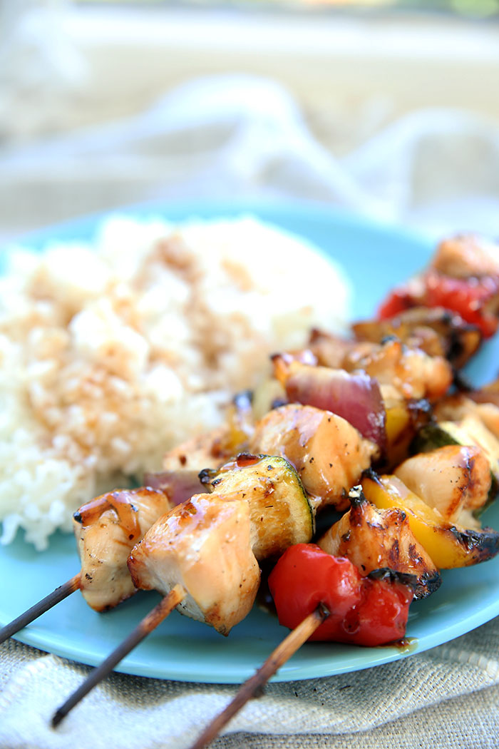 Grilled Teriyaki Kabobs in a stack of three on a blue plate with delicious coconut rice on the side. The edges of the bamboo sticks are burnt but the kabobs look delicious and were grilled to perfection.