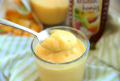 Orange smoothie looking drink in a clear glass cup with a bottle of honey and a blender and frozen mano