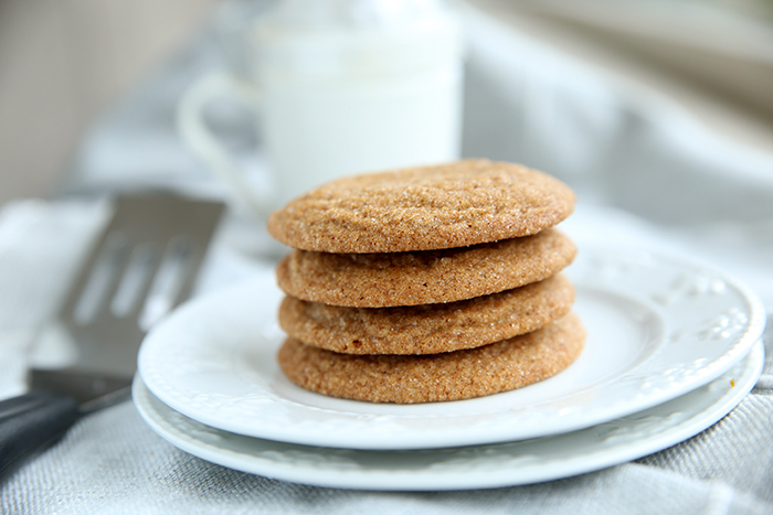 four chewy gingersnap cookies stacked on top of each other on a small white plate. There is a cookie spatula next to the plate an a white mug in the background