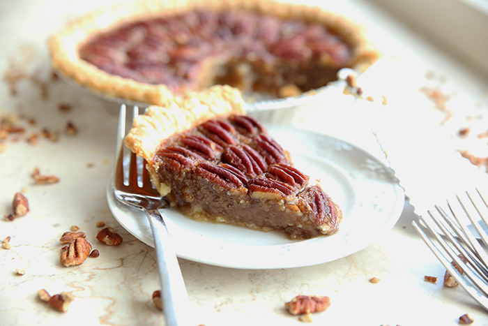 A small white plate with a slice of pecan pie on it and a fork, there is a pecan pie in the background of the picture