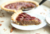 A small white plate with a slice of pecan pie on it, you can see the rest of the pecan pie in the background and a few more plates to the right