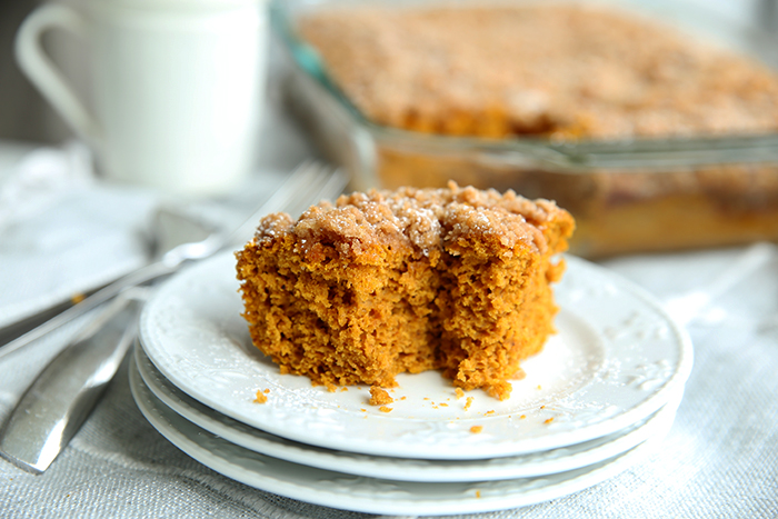A piece of Pumpkin Coffee Crumb Cake that has a bite out of it, showing the crumbs and stacked on three white plates with the rest of the cake in the background in a glass pan.