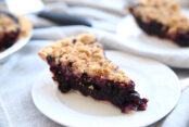 a slice of blueberry pie with a crumble topping on a white plate. A second slice is in the background and you can also see a corner of the pie pan the slices came out of.
