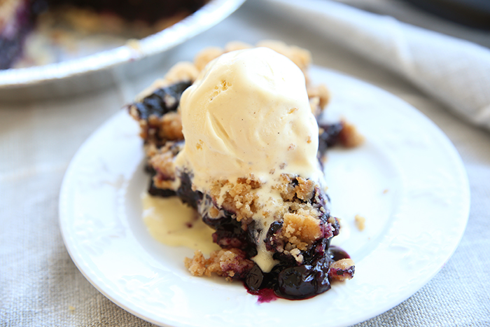 slice of blueberry crumble pie with a scoop of vanilla ice cream on top that is starting to melt. It is on a white plate. In the background you can see the corner of the pie pan with the rest of the pie in it. 