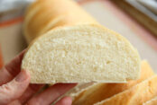 A hand holding a slice of french bread, you can see the rest of the loaf in the background.
