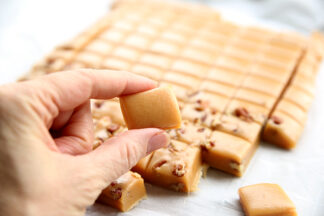 A slab of caramel cut into one inch pieces. A hand is picking up one of the pieces to eat.