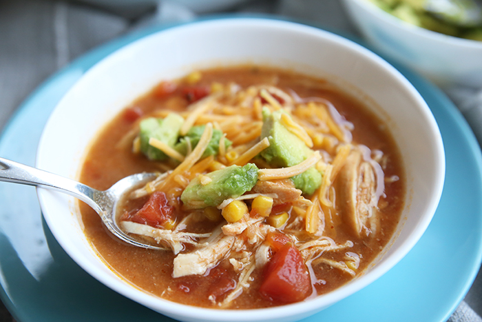 A white bowl full of chicken tortilla soup with a spoon in it. The soup is topped with grated cheese and pieces of avocado.