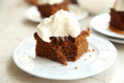 A piece of gingerbread cake with cool whip on top sitting on a white plate. Someone has taken the first bite out. In the background, you can see other pieces ready to be served.