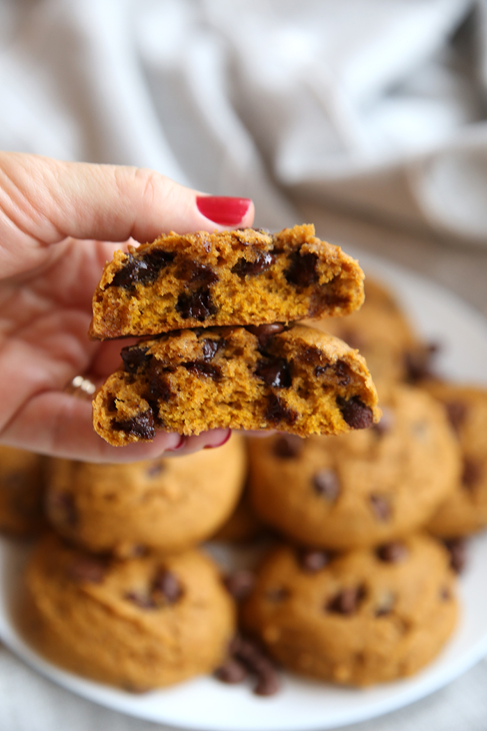 a pumpkin chocolate chip cookie that has been broken in half, it is being held in someone's hand. In the background you can see a plate of cookies