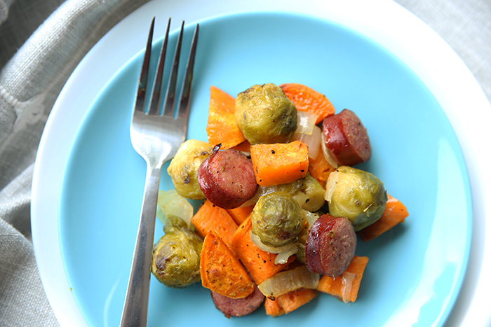 A blue plate with a scoop of chopped Kielbasa, sweet potatoes and brussels sprouts with a fork sitting on top of the plate on the leftside.