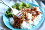 A blue plate with a fork holding a bite of Slow Cooker Teriyaki Chicken served on a bed of rice with broccoli.