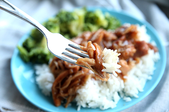 A blue plate with a fork holding a bite of Slow Cooker Teriyaki Chicken served on a bed of rice with broccoli.