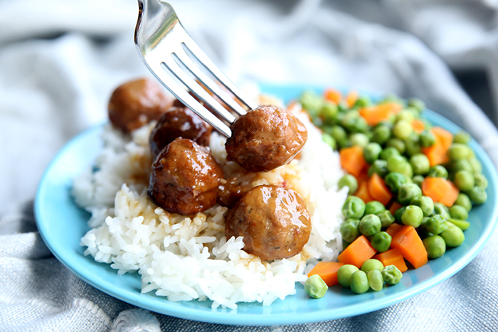 A blue plate sitting on top of a table cloth with peas and carrots and teriyaki meatballs topped on jasmine rice