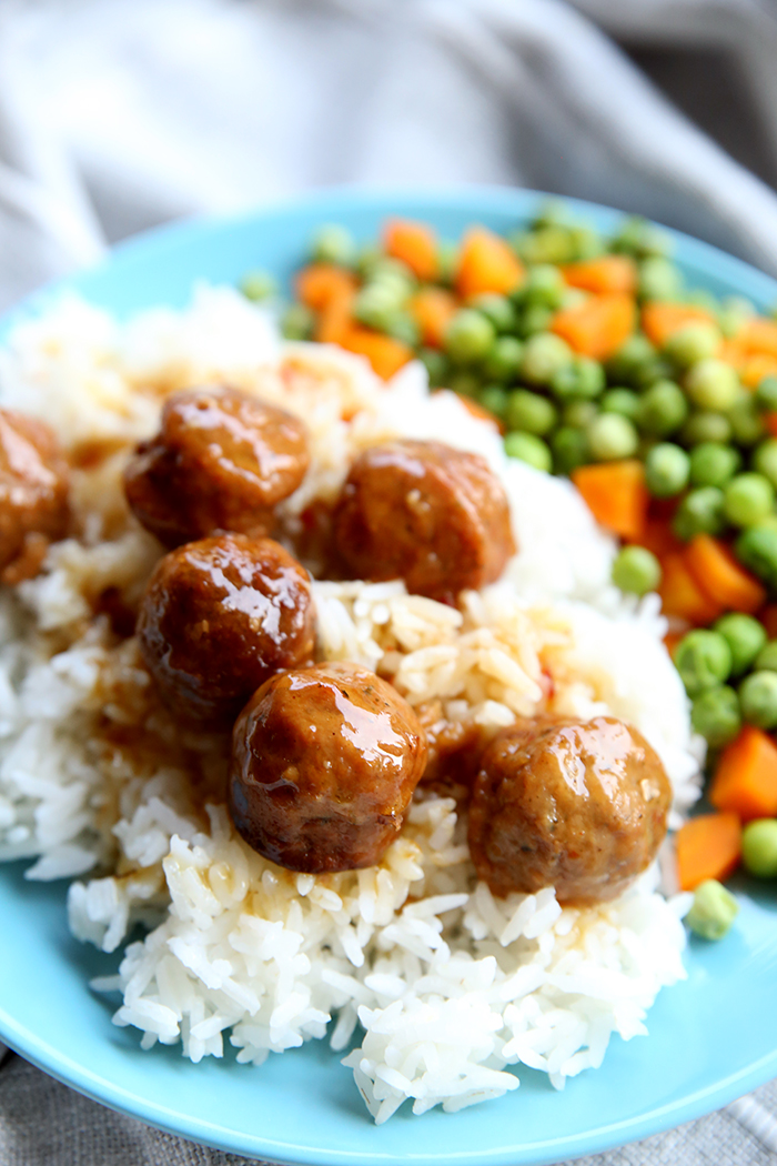 Teriyaki meatballs topped on a scoop of rice with carrots and peas on the side, all on blue plate sitting on top of a table cloth