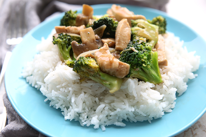 A fork is on the left side of a blue plate, sitting on a grey napkin. The blue plate has white rice on it topped with the broccoli chicken stir fry.