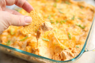 a glass dish full of buffalo chicken dip. there is a chip being dipped in and you can see the cheese pull