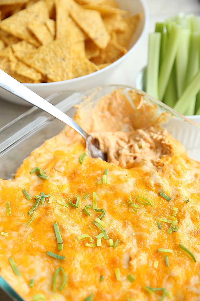 a dish of buffalo chicken dip with a spoon in it for serving. There is a bowl of corn chips in the background and also a small dish of celery slices in the background