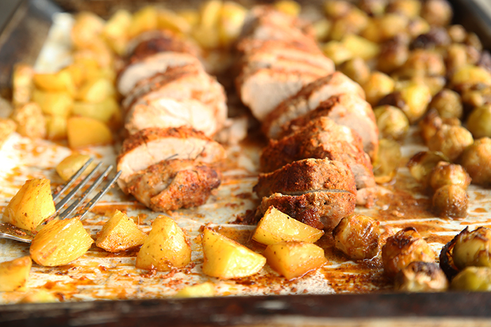 A sheet pan with sliced pork tenderloin surrounded by roasted veggies with a partial picture of a fork in the left bottom corner with a potato slice on top of it.