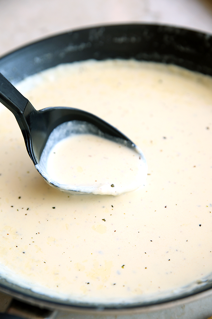 A skillet full of homemade Alfredo sauce with a serving spoon above the skillet with a scoop of sauce in it.