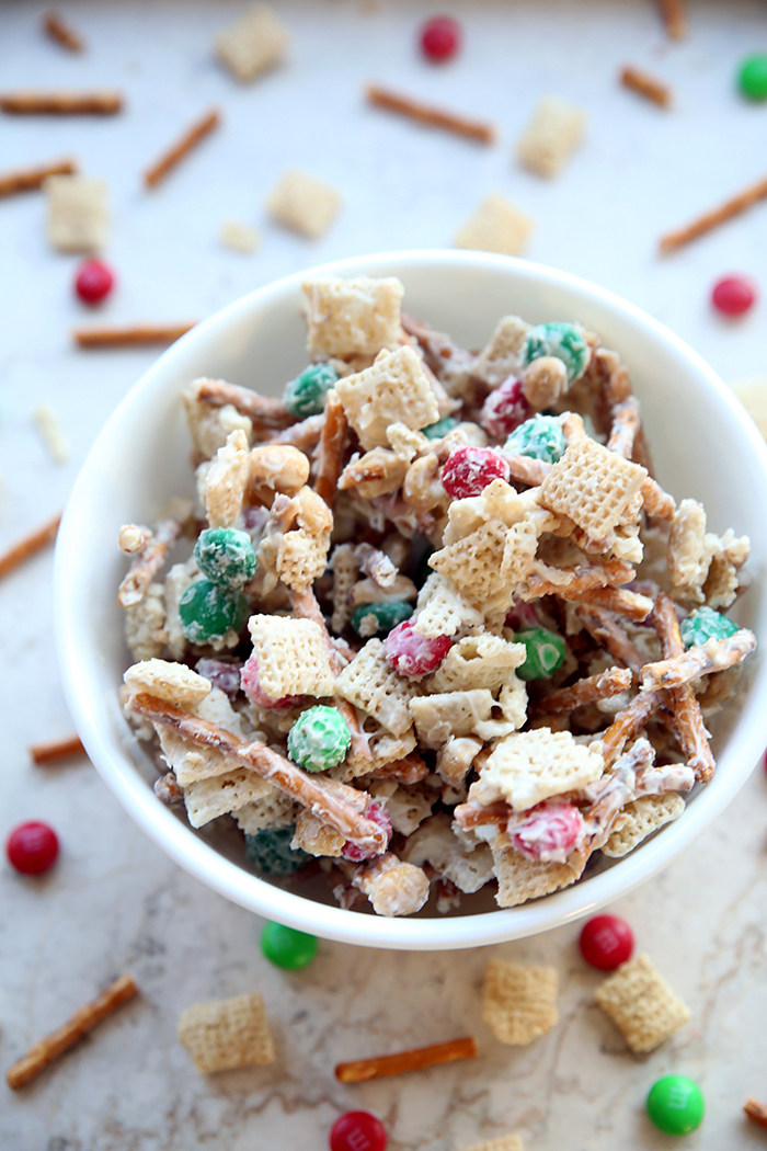 a white bowl of chex mix that has pretzels, chex cereal, peanuts and m and m's in it. it is all covered in white chocolate and there is also some scattered around the bowl