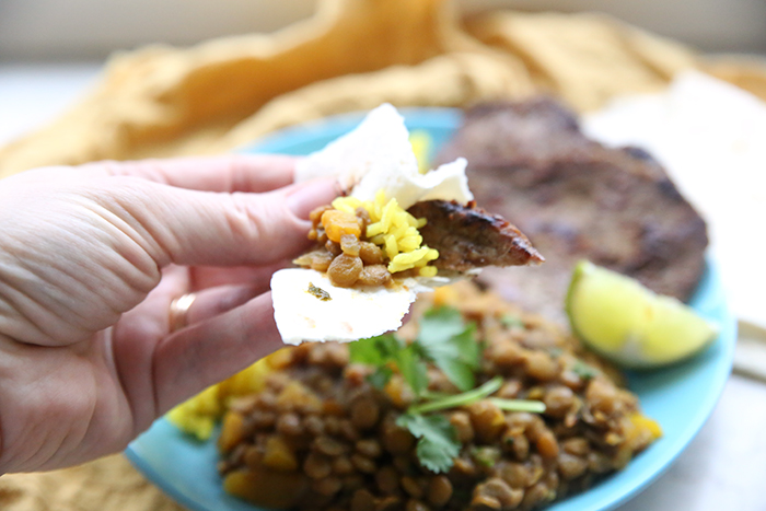 a plate of carne asada in the background with a hand holding a piece of it wrapped in a tortilla and topped with ecuadorian lentils and yellow rice. 