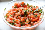 A glass bowl filled to the top with Pico de Gallo with the handle of a spoon sticking out of it.