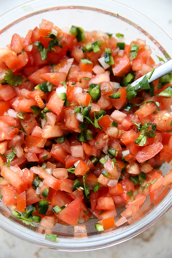 A glass bowl full of Copycat Chipotle Pico de Gallo. There's a spoon handle sticking out of the salsa on the right side.