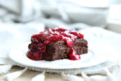 white plate with a slice of chocolate dessert that has berry glazed drizzled over it. It is sitting on a grey table cloth