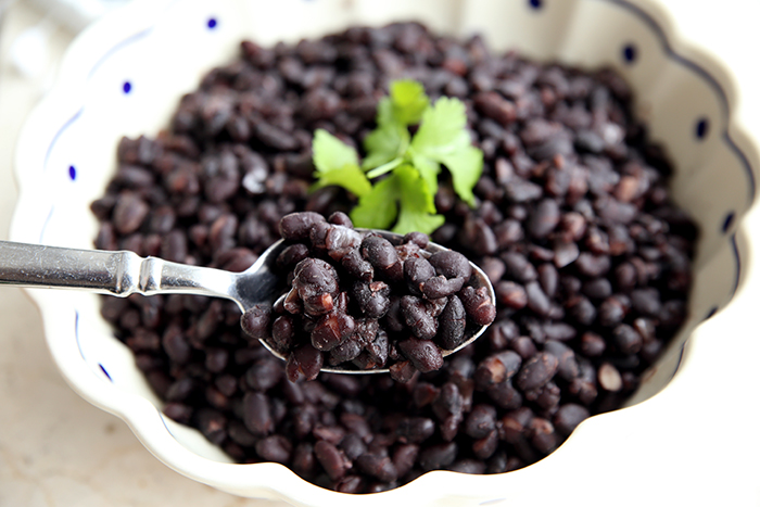 A white scalloped bowl holding instant pot black beans with a silver spoon holding a bite of beans. Cilantro garnished on top. 
