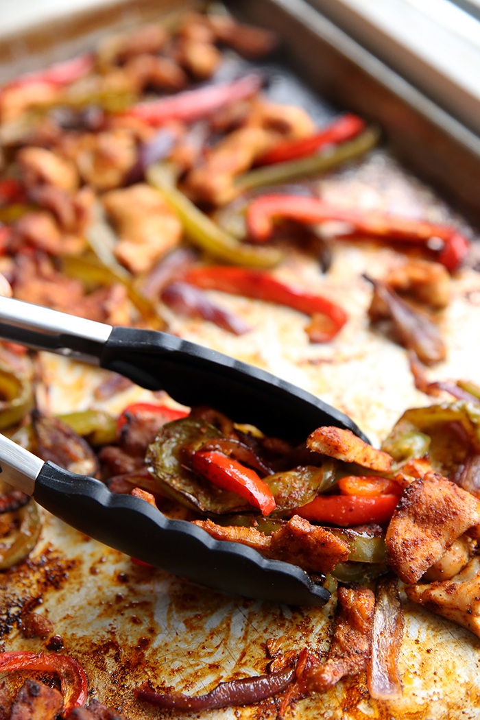 A partial picture of a sheet pan topped with chicken and bell pepper fajitas. There's a metal tong with black rubber tips diagonally placed to pick up a scoop of the fajitas