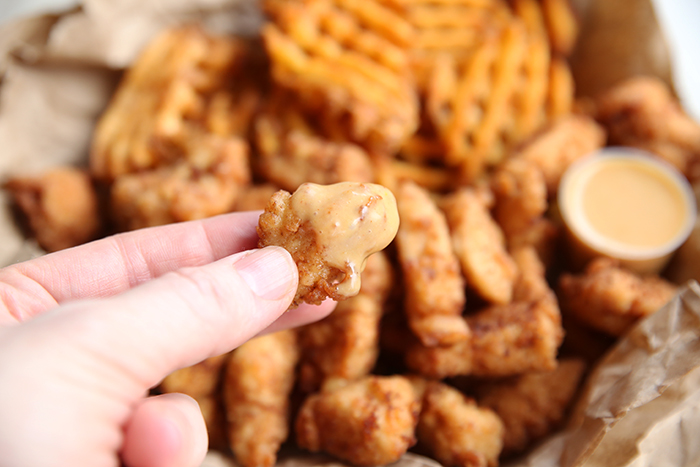 A piece of chicken dipped in chick-fil-a sauce, a person is holding it in their hand and you can see a basket of chicken in the back ground