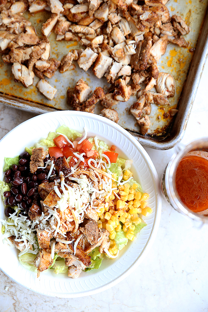 A white bowl with lettuce topped with Copycat Chipotle Chicken, black beans, Pico de Gallo, corn and cheese. On the side of the bowl is chipotle chicken dressing in a small cup. Above the bowl is a sheet pan with chopped chipotle chicken.