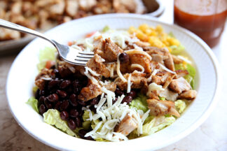 A white bowl of chipotle chicken with lettuce, corn, Pico de Gallo, black beans and shredded cheese. There's a fork getting ready to lift a bite out from the left side of the bowl. At the top of the photo in the background there's a clear cup of dressing and part of a sheet pan with cut up chicken.