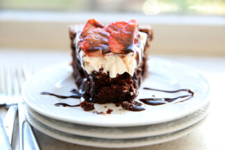 A piece of flourless fallen chocolate cake on a stack of three white plates topped with strawberries, whipped cream and chocolate sauce and a dusting of powdered sugar.