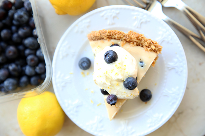In the background are a couple of lemons, two forks, and a container of blueberries. There's a white dessert plate with a slice of lemon blueberry pie topped with whipped cream and a few blueberries. On each side of the slice of pie is a single blueberry.