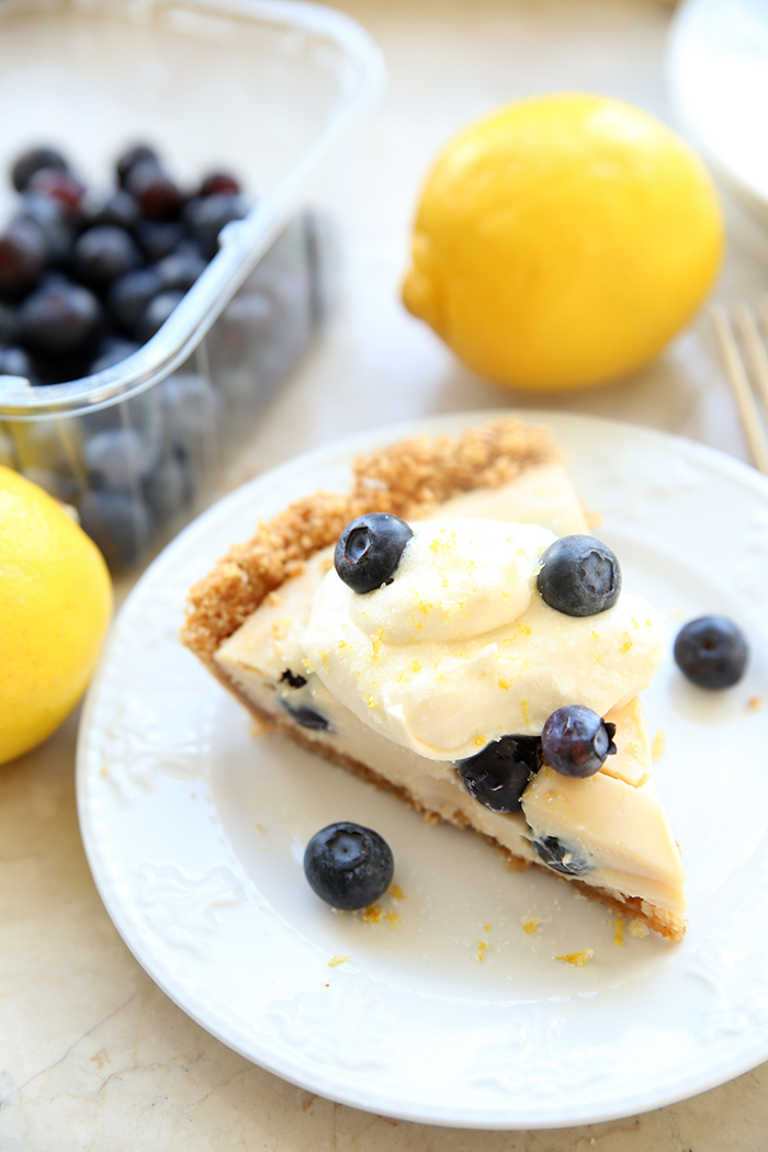 A piece of Lemon Blueberry Chiffon Pie topped with a few blueberries sitting on a small white plate with a couple of blueberries on the plate. In the background are two lemons and a plastic container of blueberries.