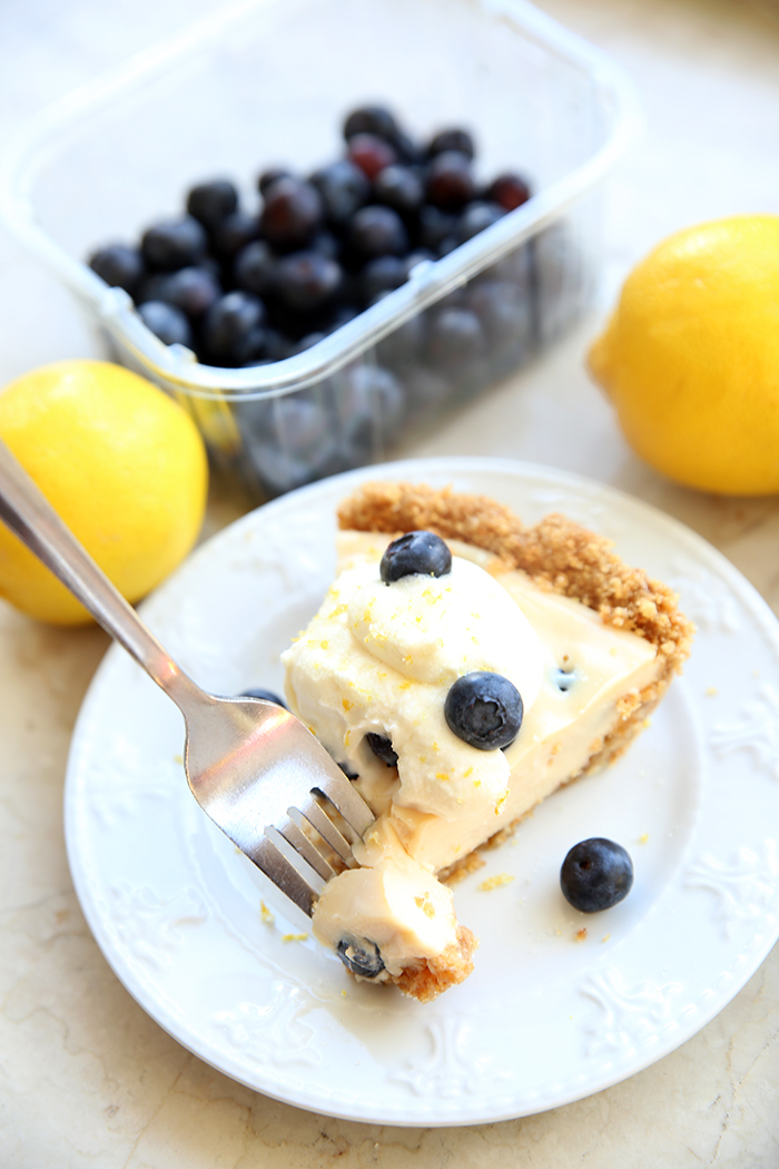 A slice of citrus chiffon pie topped with whipped cream and blueberries all on a small white plate. A fork is being held diagonally up while taking a piece out of the slice. The background has a container of blueberries and two lemons.