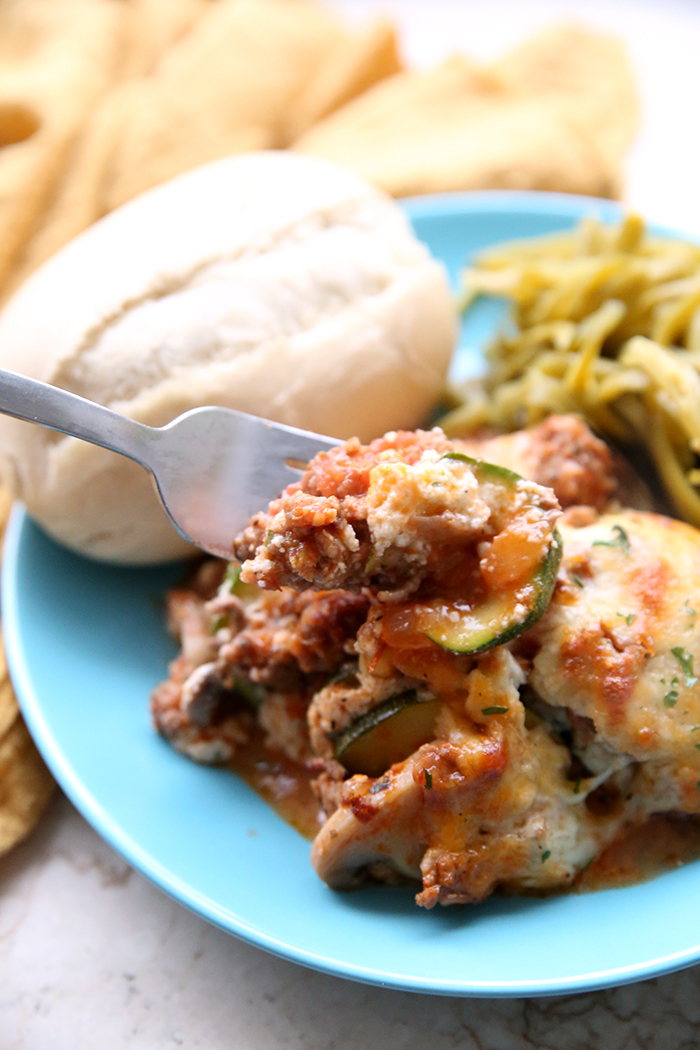 A sky blue plate with a roll, green beans and a serving of zucchini lasagna with a forkful of lasagna held above the plate. In the upper background, there's a yellow cloth napkin.
