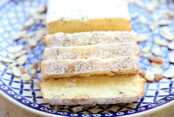 A white and dark blue patterned plate with a loaf of Almond cake dusted with powdered sugar with four slices cut out and almonds scattered around the plate.