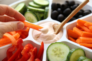 A hand holding a sliced carrot with dip on it is above a 7-compartment vegetable tray filled with carrots, bell peppers, cucumbers, olives and Fiesta Ranch Dip. In the dip is a small wooden spoon sticking out of the dip.
