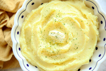 Mashed potatoes with a small scoop of butter in the middle with parsley sprinkled on top all in a white Polish potter bowl with scallops, blue trim at the top, and dark blue polkadots throughout the bowl. On the left side of the bowl is a cloth yellow napkin scrunched up.